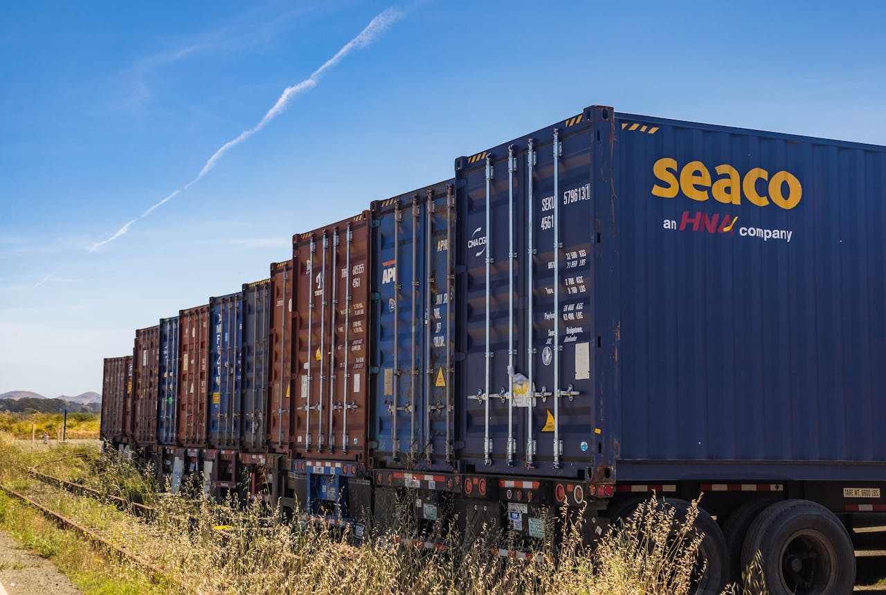 hero-img-02 Row of vibrant cargo containers on train tracks under a clear blue sky.