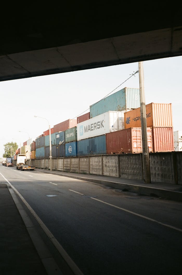 services-03 Multicolored cargo containers stacked beside a busy urban street under a clear sky.